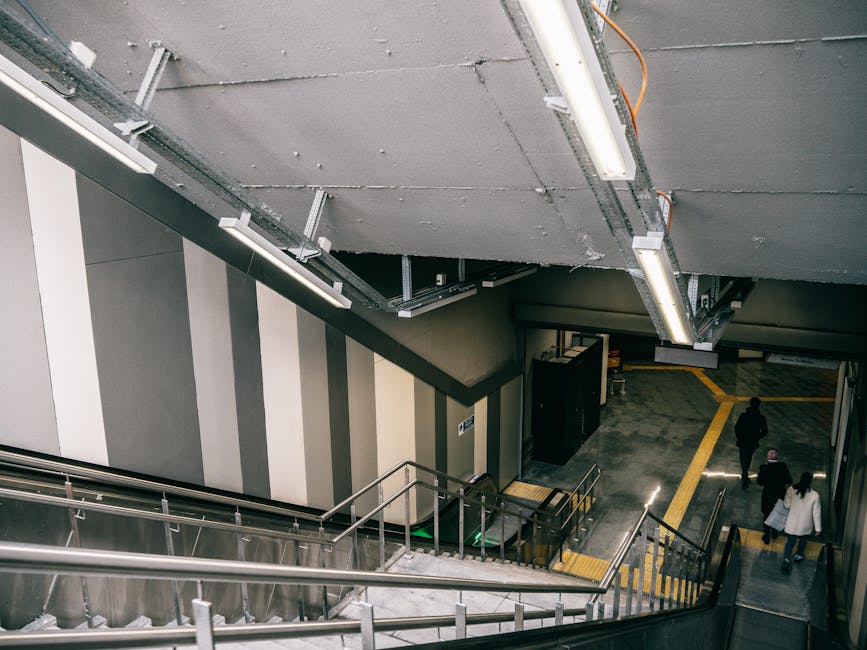 The image shows an indoor stairwell area within a public transport station, with a downward view of the staircase leading to an underground passage. The staircase has metallic railings and steps, and 