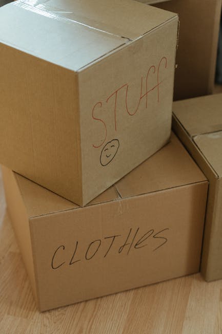 Interior view of a residential property during a house move, showing a person standing beside several cardboard boxes on a wooden floor. Some boxes are stacked, while others are positioned individuall
